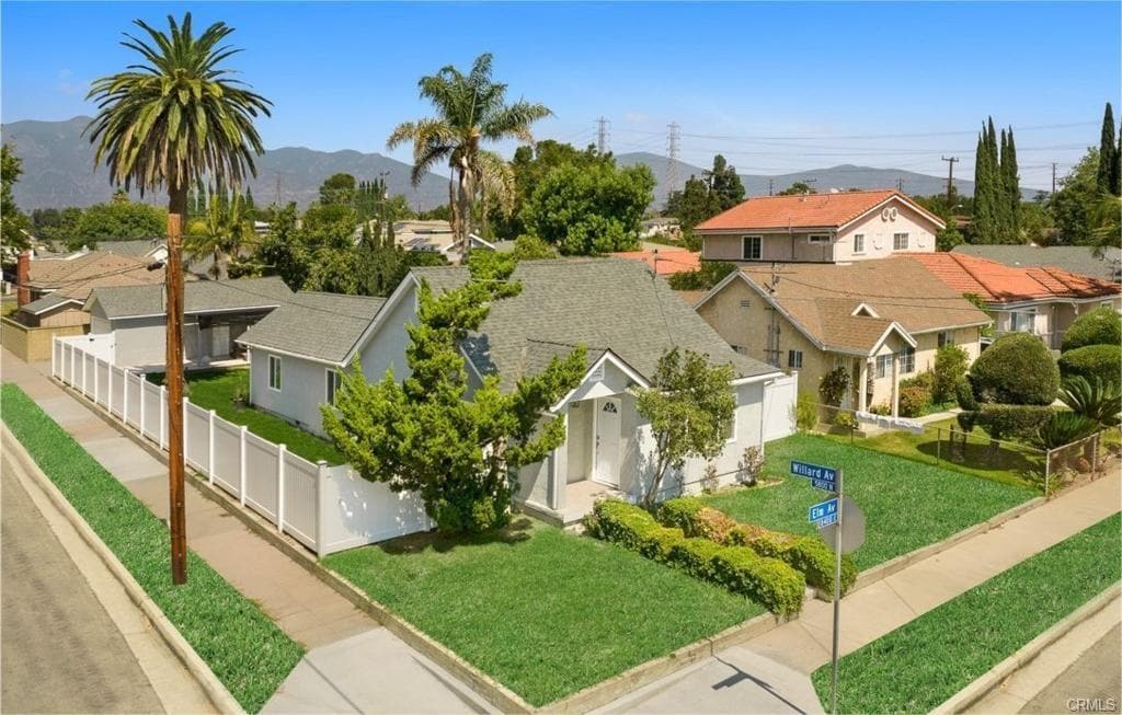 A single-family house is depicted from an aerial view, showcasing a well-maintained yard surrounded by a low white fence. Lush greenery and mature trees are visible, providing a sense of space. The neighborhood includes various homes, palm trees, and a clear blue sky.
