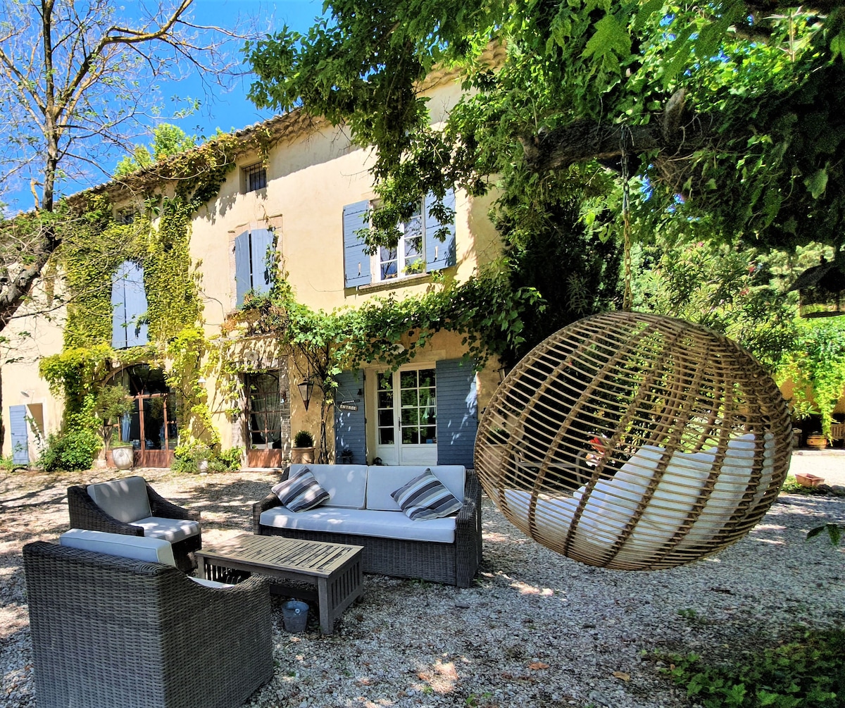 A shaded outdoor seating area features a comfortable arrangement of wicker furniture and a hanging chair, surrounded by lush greenery. Sunlight filters through the leaves, creating a serene atmosphere in front of the charming stone house with blue shutters.