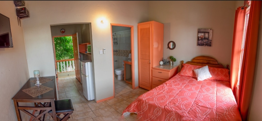 A studio apartment features a spacious bed with a decorative orange coverlet. A dining area with a small table and two chairs is situated in the room. A kitchenette is visible in the background, along with a notable open door leading to a bathroom.