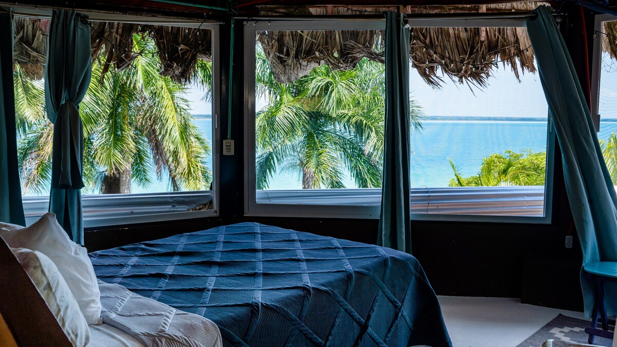A spacious sleeping area is highlighted by a large bed, covered with a deep blue quilt. Expansive windows frame views of the tropical landscape and blue water beyond, allowing natural light to fill the room.