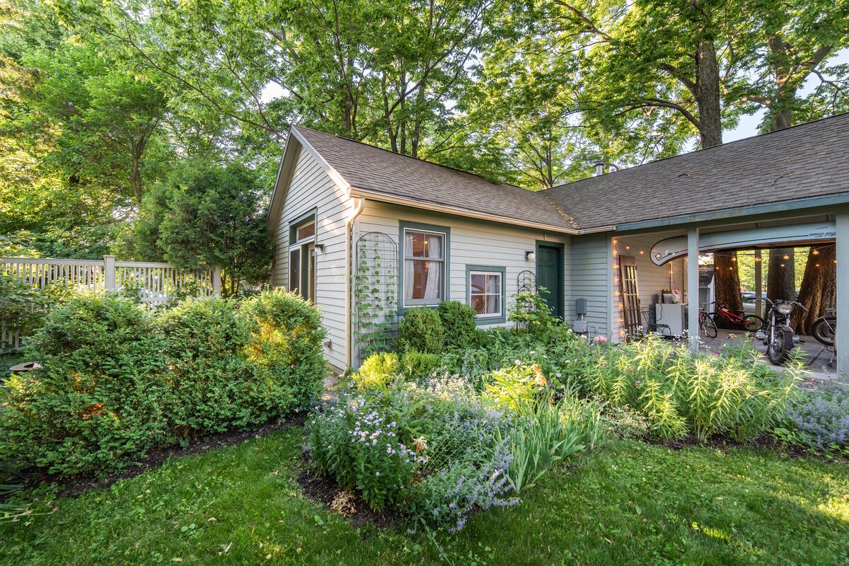A well-maintained exterior of the casita is surrounded by lush greenery and garden beds filled with various plants. The entrance features a modest porch with a window above, while a carport is visible on the side, partially shaded by the surrounding trees.