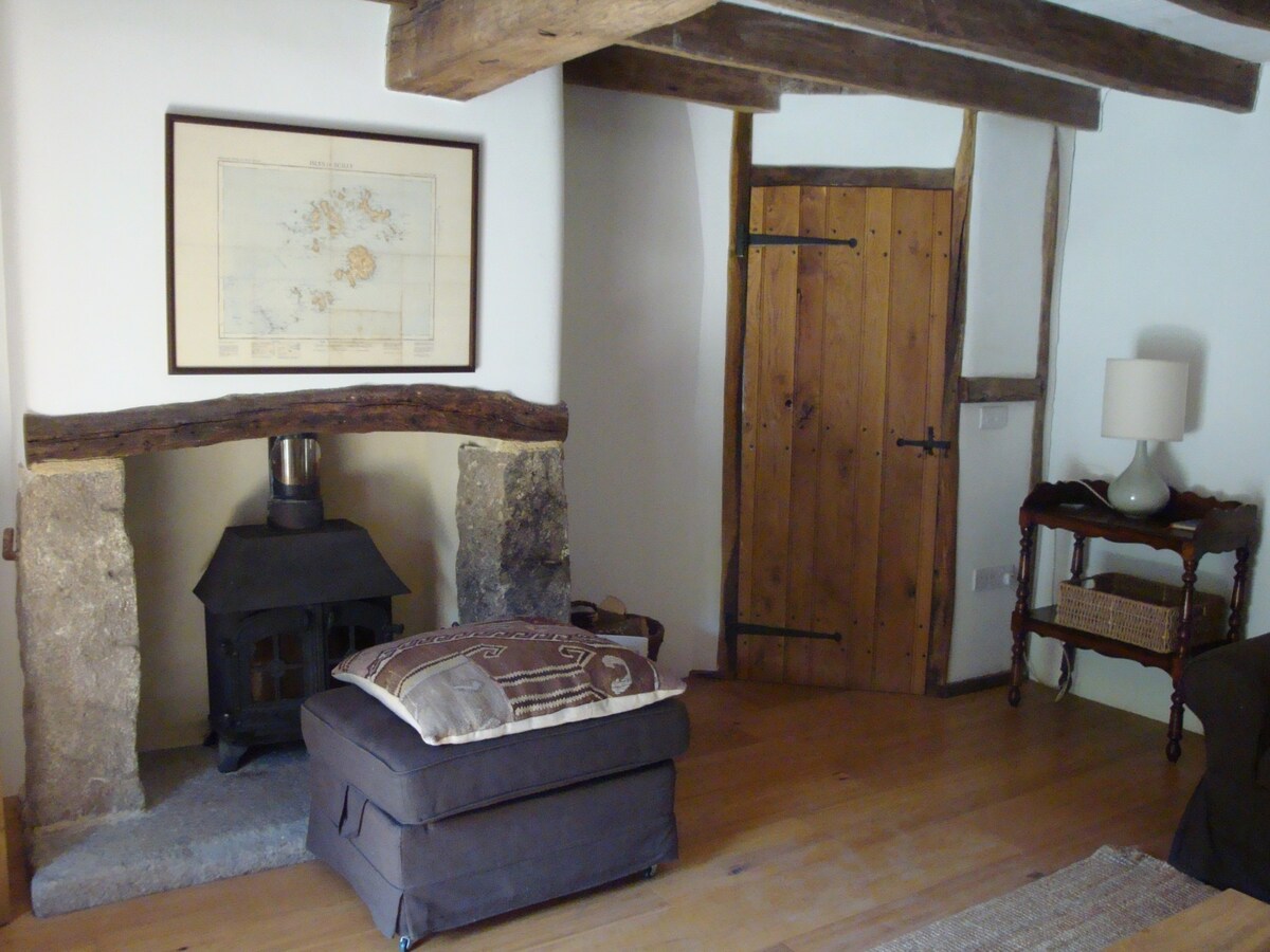 The sitting room features exposed wooden beams and a rustic charm. A wood burner is positioned on a stone base, complemented by a decorative ottoman. Warm wooden flooring extends throughout, while a wooden door and a framed map add character to the space.