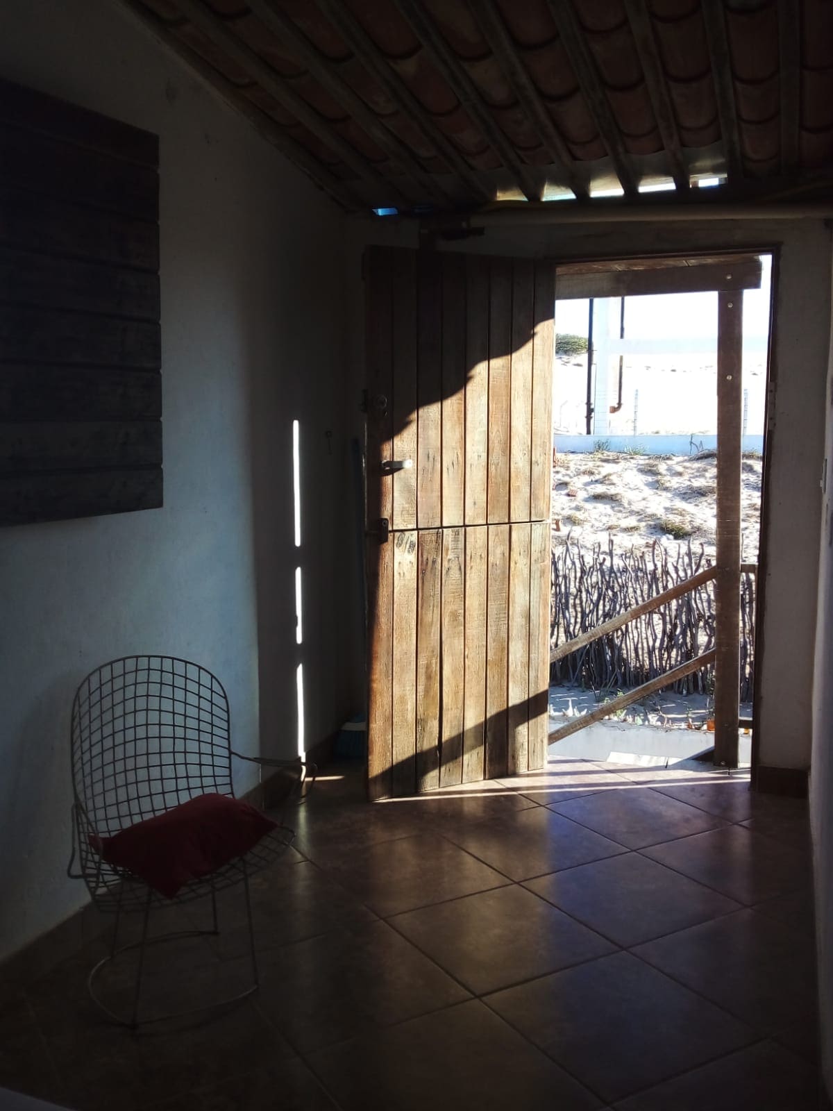 Natural light fills a welcoming entryway, where a wooden door stands partially open. A textured wire chair is positioned nearby, complemented by a red cushion. Beyond the threshold, a view of a rustic outdoor area is visible.