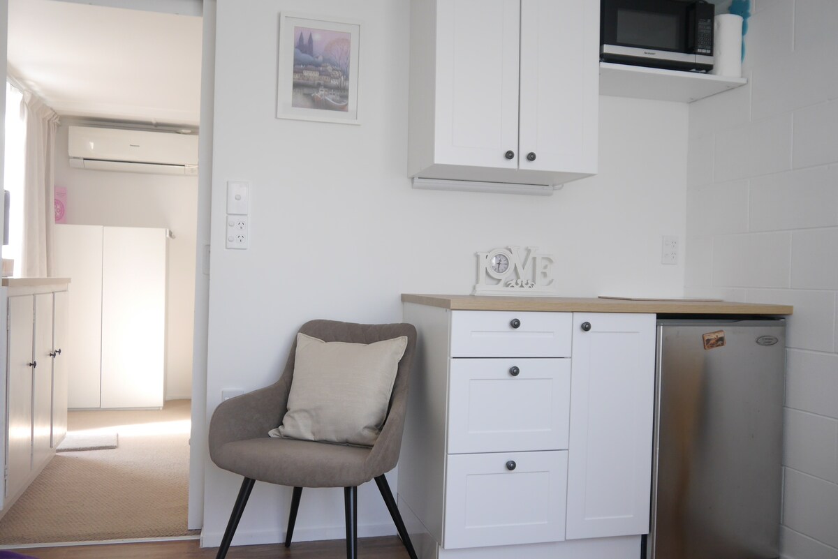 A kitchenette area showcases a compact refrigerator, microwave, and cabinetry, providing essential appliances for food preparation. A cozy chair sits beside a small table, while light streams in from a nearby window, illuminating the neutral-toned walls and soft furnishings.