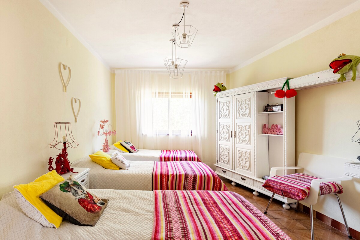 A bright bedroom is presented with three beds, each adorned with colorful striped blankets. A white cabinet with ornate detailing and playful decor is located beside a window, which allows soft light to fill the space. Decorative lamps add a charming touch.