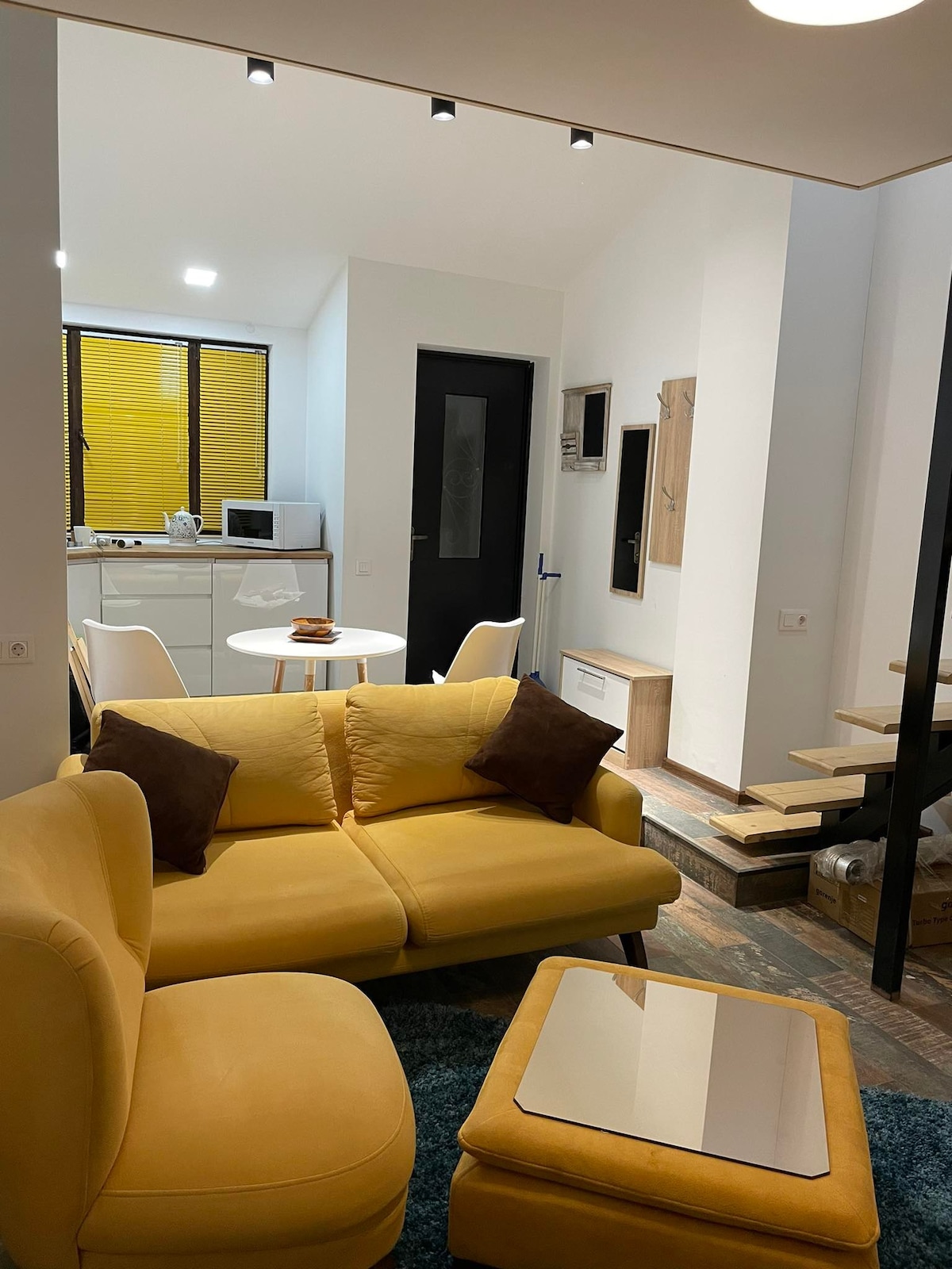 A compact living area features a yellow sofa adorned with two brown cushions. A round white dining table with two chairs is positioned nearby. Bright yellow window blinds allow filtered light into the space. A staircase is visible, leading to an upper level.