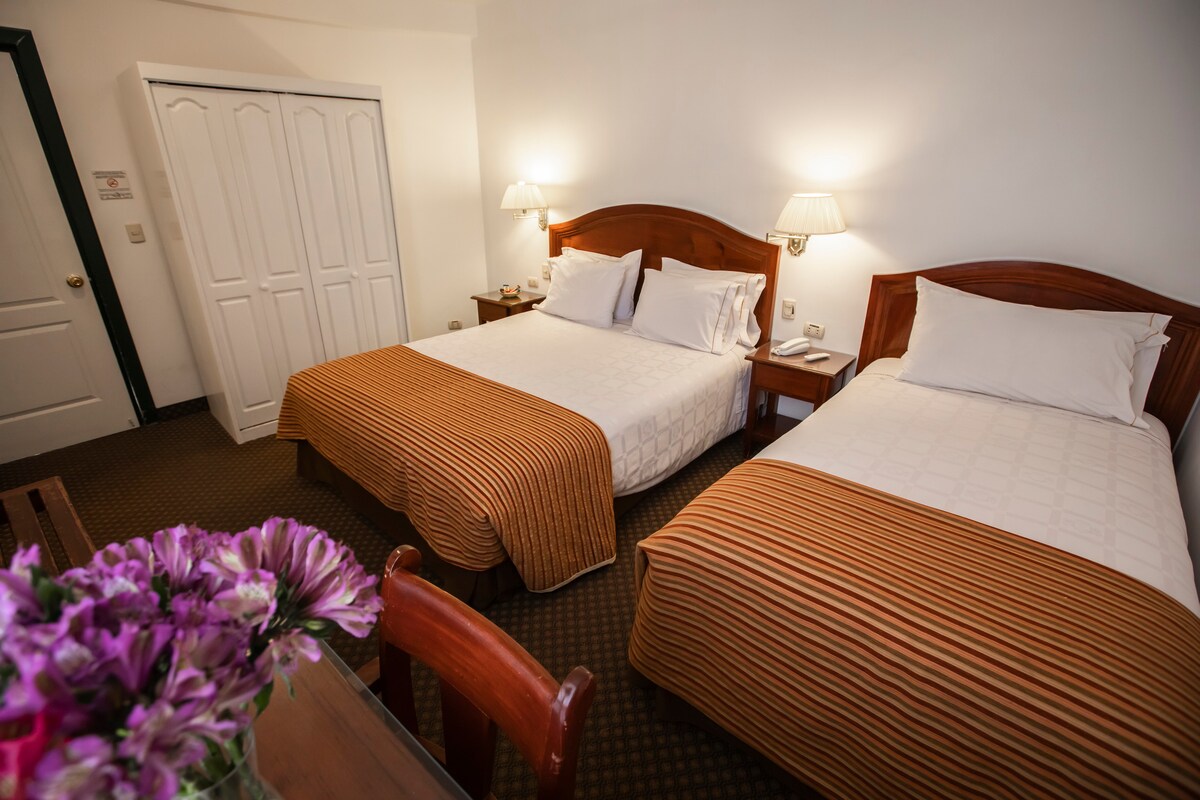 A guest room is presented with two spacious beds, each topped with striped brown and cream blankets. A wooden dresser is visible, along with a bouquet of flowers on the table. Soft lighting emanates from wall sconces near the beds, creating a cozy ambiance.