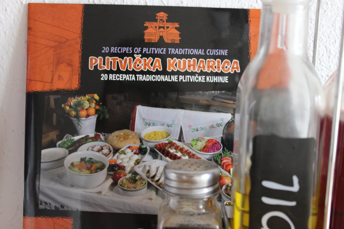 A cookbook titled 'Plitvička Kuharica' is displayed, featuring an array of traditional dishes from the Plitvice region. A colorful spread of food is visible, accompanied by fresh ingredients. Various bottles of cooking oils and ingredients are positioned alongside the book, enhancing the home cooking theme.