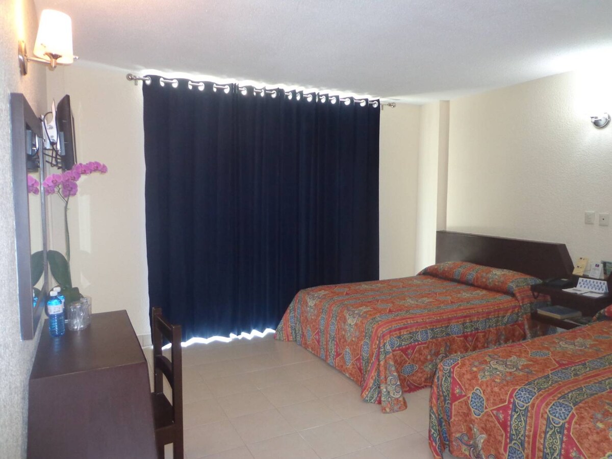 The room features two beds adorned with colorful patterned bedding, complemented by a wooden headboard. A small desk and chair are positioned against the wall. Dark curtains cover a window, and a television is mounted above a cabinet with additional furnishings.