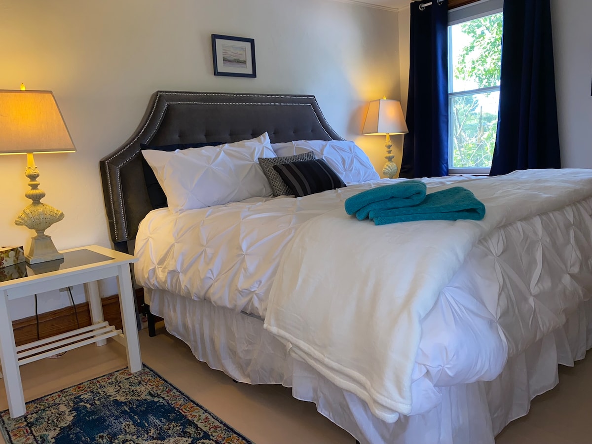 A spacious bedroom is highlighted by a plush bed covered in crisp white linens. Flanking the bed are two bedside lamps providing soft illumination. A large window with dark curtains allows natural light to filter in, while a cozy rug adds texture to the floor.