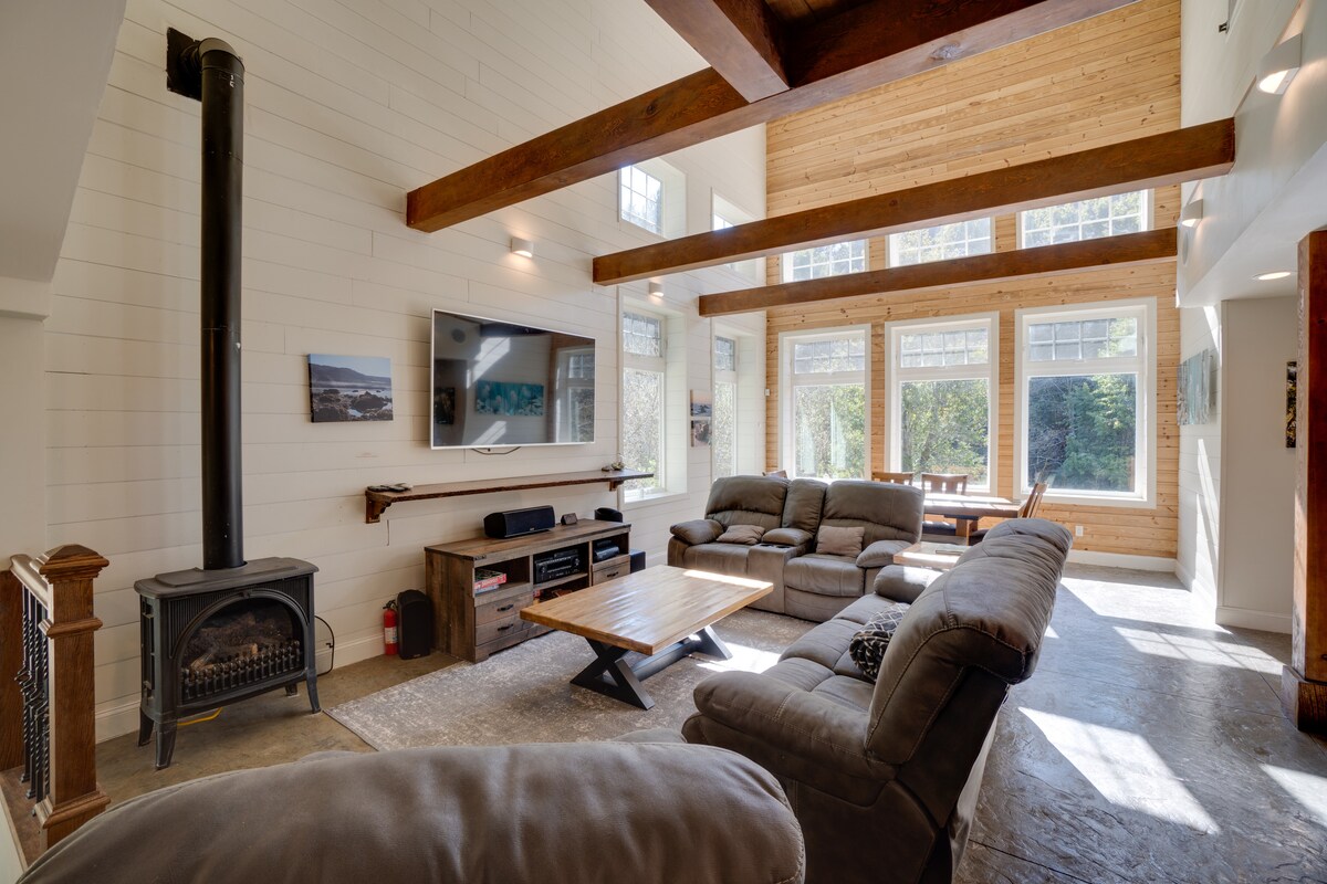 A spacious great room features high ceilings with exposed wooden beams and large windows providing natural light. Comfortable seating is arranged around a coffee table, with a sleek entertainment unit and a cozy wood stove accentuating the welcoming atmosphere.