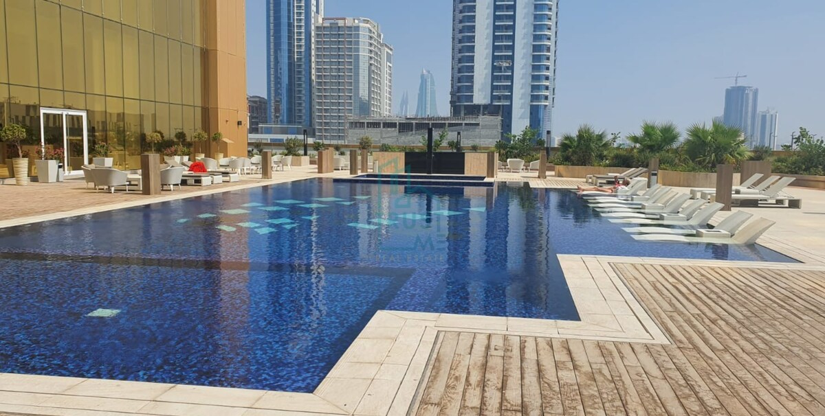 A spacious outdoor pool is surrounded by a wooden deck and lounge chairs. The serene blue water reflects the clear sky and nearby skyscrapers, creating an inviting area for relaxation. Lush greenery and contemporary seating enhance the modern aesthetic of the poolside.