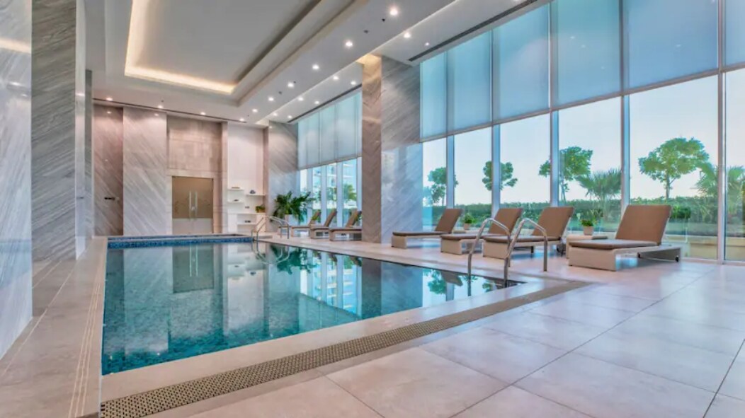 An indoor swimming pool area is surrounded by large glass windows, allowing natural light to fill the space. Lounge chairs are positioned around the pool, with a sleek tile design complementing the modern aesthetic. Lush greenery is visible through the windows, creating an inviting atmosphere.