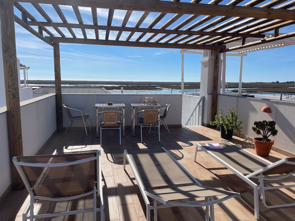 A spacious terrace is featured with a wooden pergola, providing shade for the dining area and lounge chairs. A table with chairs sits adjacent to potted plants, while panoramic views of the tranquil surroundings can be seen in the background.