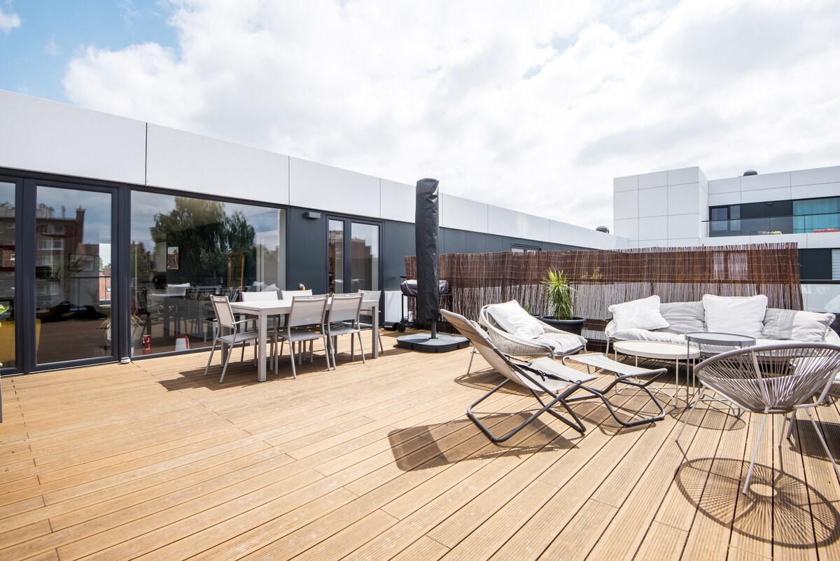 The spacious terrace is highlighted by a wooden deck, offering a large table with six chairs and four lounge chairs. A tall privacy screen surrounds part of the area, while a vertical garden adds greenery. A barbecue grill is present, enhancing the outdoor setting.