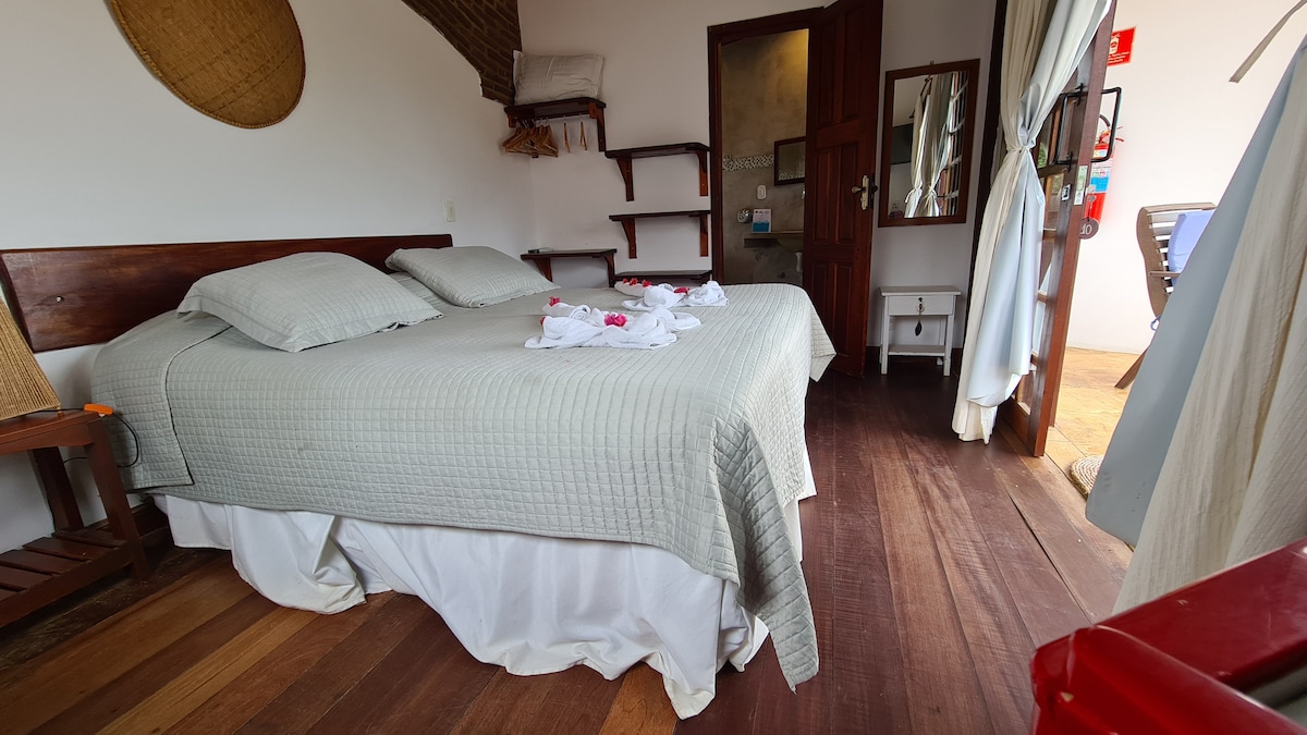 A spacious bed with light grey bedding is positioned on wooden flooring, complemented by soft towels arranged on top. Nearby, a doorway leads to a well-lit area, while shelves are mounted on the wall above a small wooden bench.