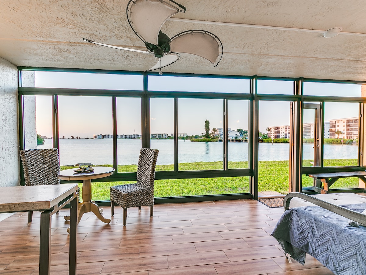 A bright Florida room features large glass windows offering expansive views of the riverfront. Two wicker chairs are positioned at a small table, inviting relaxation. Wooden flooring enhances the spacious and open layout, where light fills the area, creating a serene atmosphere.