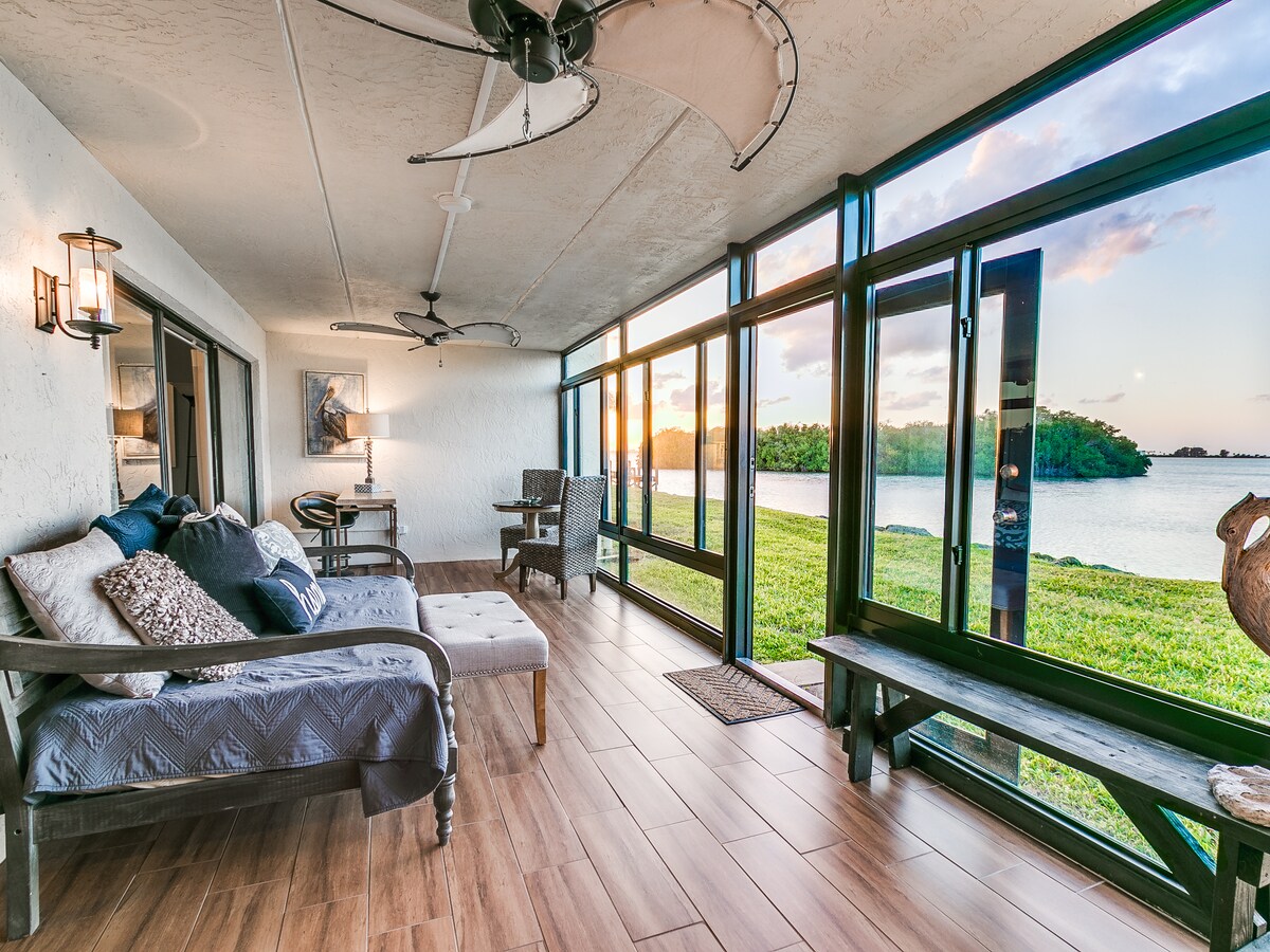 The Florida room features large glass windows offering expansive views of the river. Comfortable seating is arranged with soft cushions, complemented by a dining area for two. Natural light fills the space as sunset hues illuminate the interior.