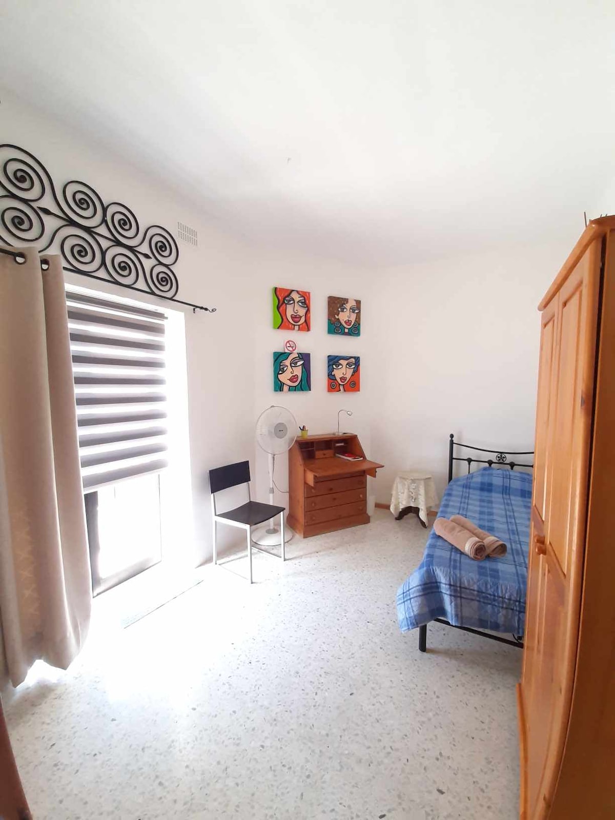 A well-lit bedroom features a single bed with a blue blanket, accompanied by a wooden wardrobe and a small writing desk. A black chair and a fan are present. Natural light enters through a window adorned with modern blinds, enhancing the room's airy feel.