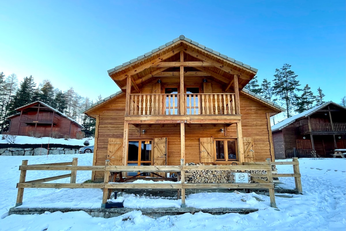The chalet is constructed from natural wood, featuring a prominent balcony and timber accents. A spacious porch is visible, surrounded by snow-covered ground. The structure stands among trees, with additional wooden buildings in the background, enhancing the peaceful outdoor setting.