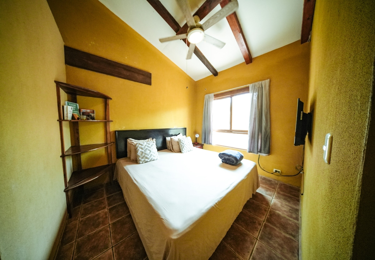 A spacious bedroom features a king bed with neutral bedding, complemented by decorative pillows. A wooden shelf holds various items. Light enters through a window covered by sheer curtains, and a ceiling fan adds to the comfort of the room.