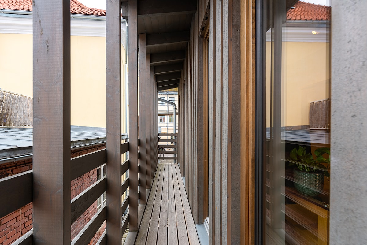 A narrow wooden balcony extends outside, flanked by wooden panels on either side. The floor consists of laid timber planks, and large glass doors reveal views of the interior space. Natural light filters in, enhancing the warm tones of the wood.
