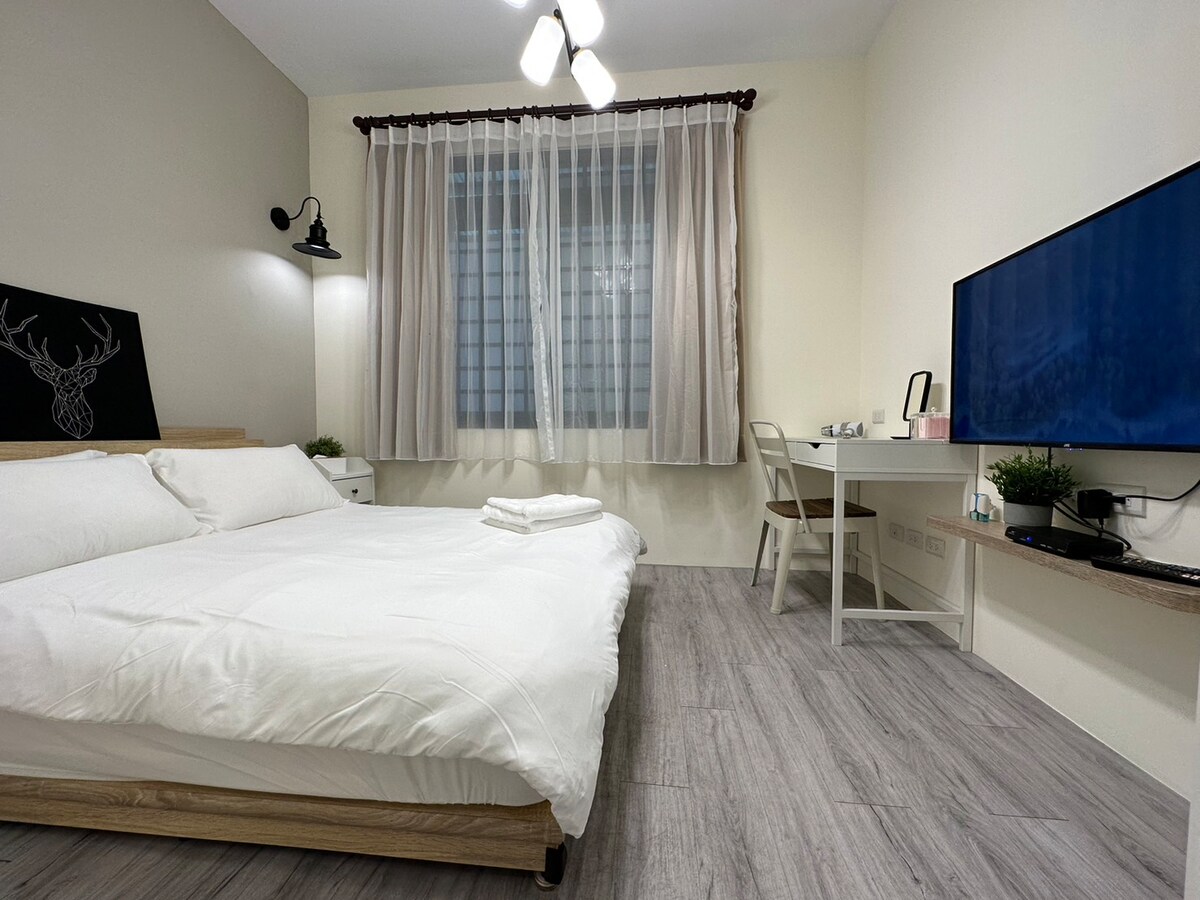 A cozy sleeping area features a spacious bed with white linens, complemented by an adjacent desk and chair. Soft natural light filters through sheer curtains, and a TV is mounted on the wall. A small green plant adds a touch of decor to the room.