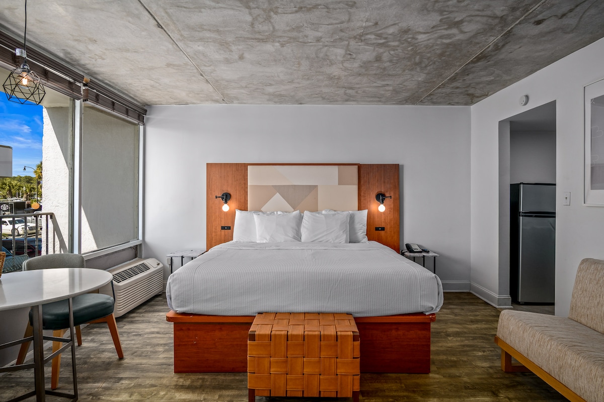 A modern hotel room features a spacious bed with a graphic patterned headboard. Two bedside lamps provide soft lighting. A sitting area includes a round table and chairs. A mini-fridge and microwave are present, complementing the neutral tones of the decor.