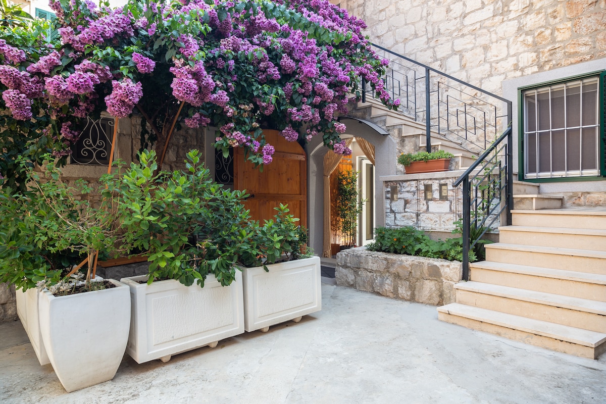 A serene front garden is adorned with vibrant purple flowers cascading over the entrance. The stone wall creates a charming backdrop, while decorative planters filled with greenery add a touch of nature. Steps lead to the apartment entrance, enhancing accessibility.