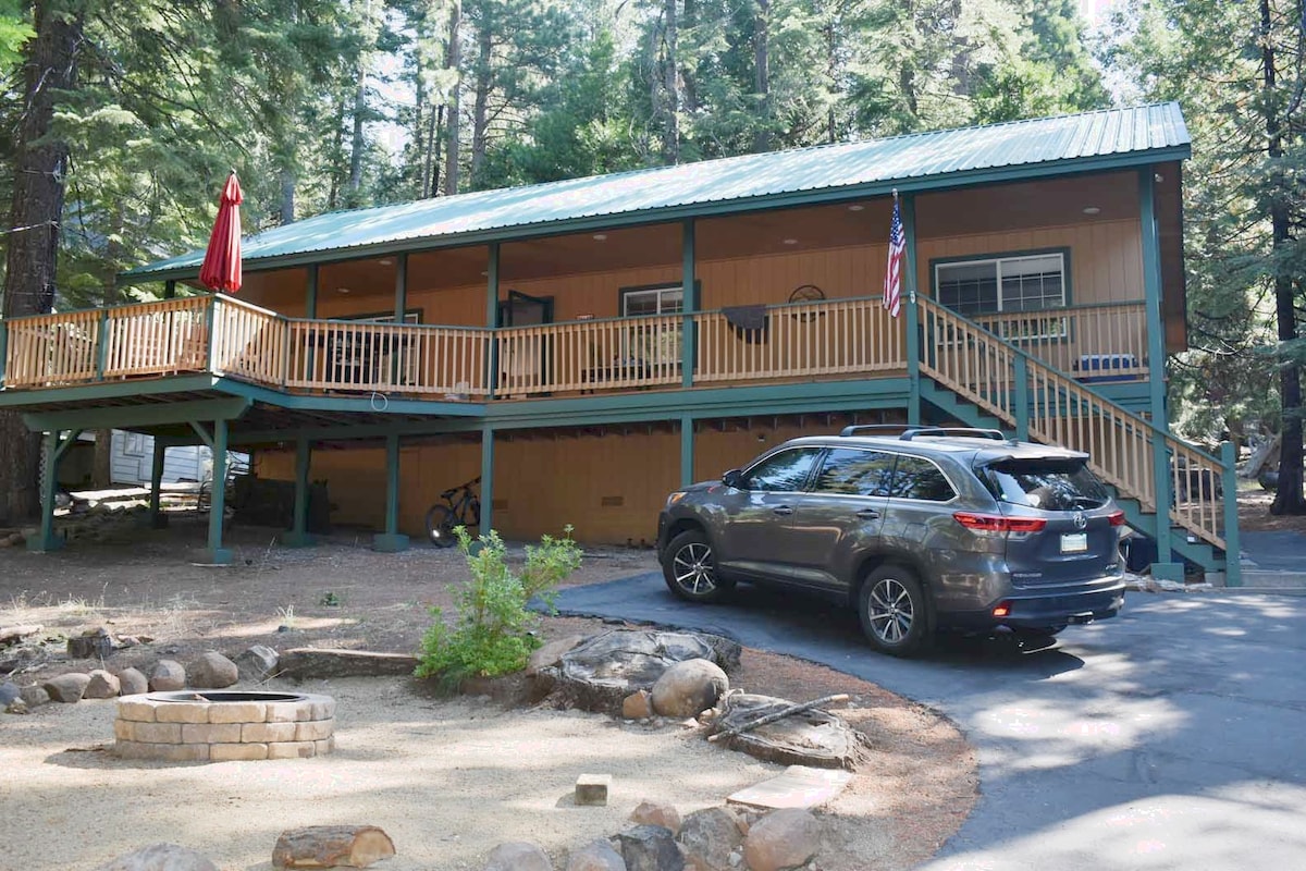 A welcoming home is set among tall trees, featuring a wooden deck with a red umbrella. A gray SUV is parked on the driveway, which leads to a stone fire pit area enhanced by natural landscaping.