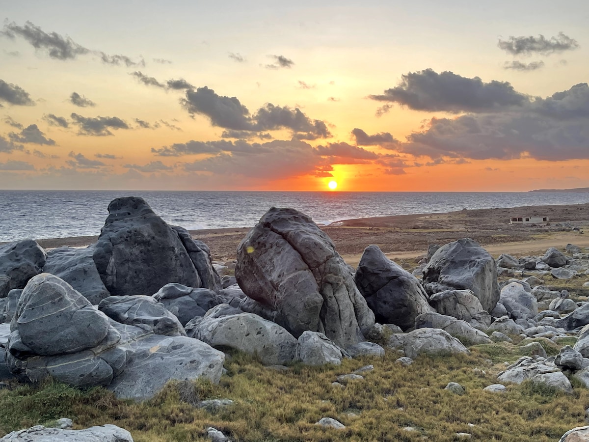 A serene landscape features large, textured boulders in the foreground, set against a dramatic sunset over the ocean. Soft clouds scatter across the sky, and gentle waves can be seen meeting the shoreline in the distance.