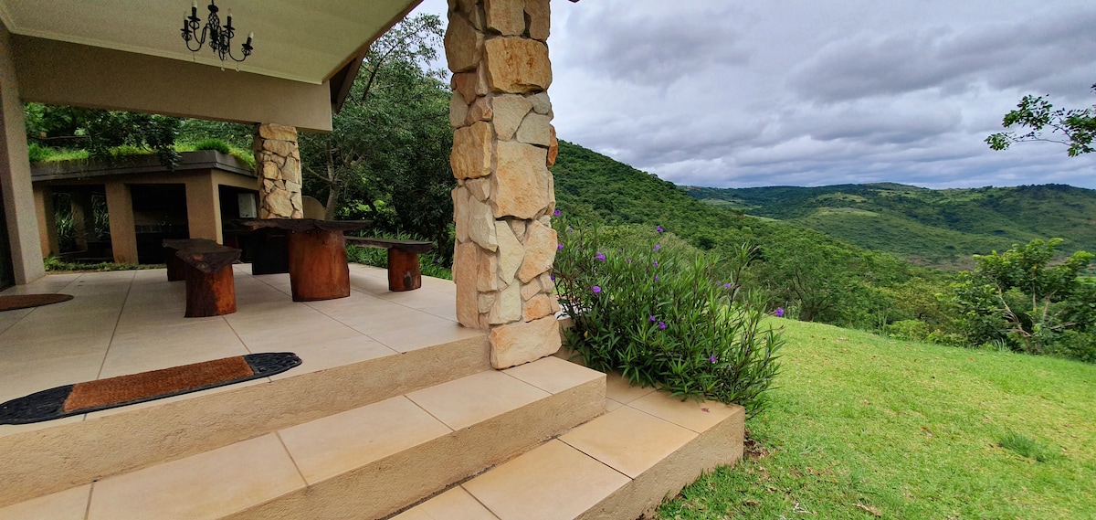 A stone porch leads to a spacious outdoor seating area, featuring a large wooden table surrounded by natural greenery. A sweeping view of rolling hills and lush vegetation is visible in the background, enhancing the serene outdoor setting.
