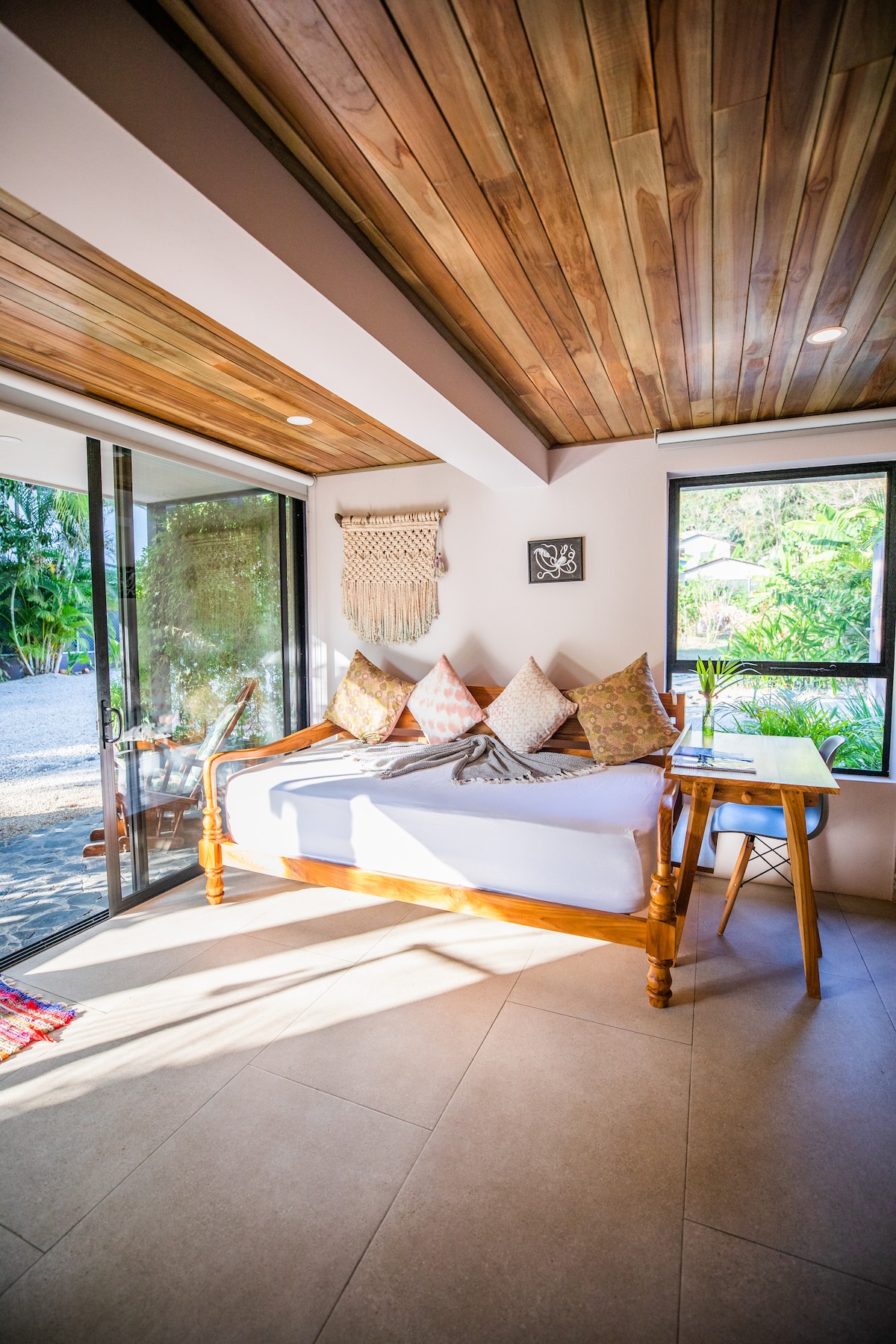 A bright sleeping area features a wooden daybed adorned with decorative pillows. Natural light floods in through large glass doors, revealing views of the lush garden outside. A small desk is positioned beside the daybed, with a decorative lamp that adds an inviting touch.