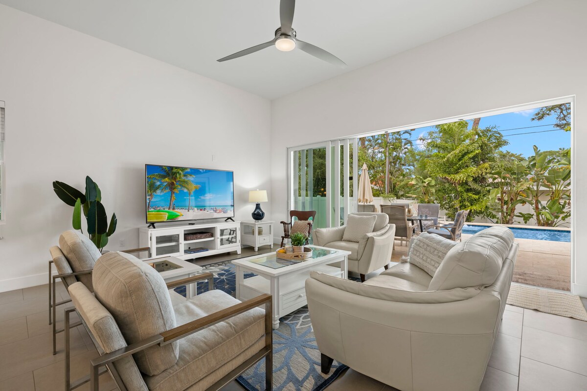 A modern living area features a comfortable seating arrangement with light-colored chairs and a sofa. A flat-screen TV is mounted on the wall, showcasing a tropical scene. Large glass doors open to reveal a view of palm trees and a private pool area.