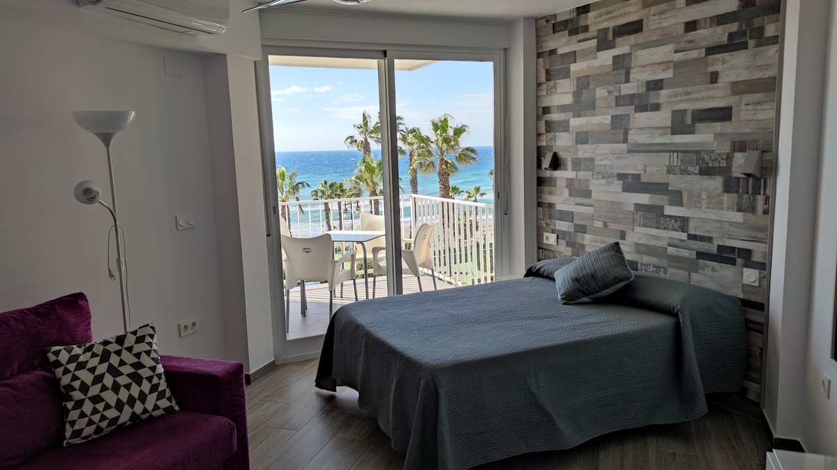 A cozy studio layout features a spacious bed covered in a neutral blanket. A large window offers stunning views of the sea and palm trees. A comfortable purple sofa with geometric cushions is positioned nearby. Sliding doors lead to a terrace with outdoor seating.