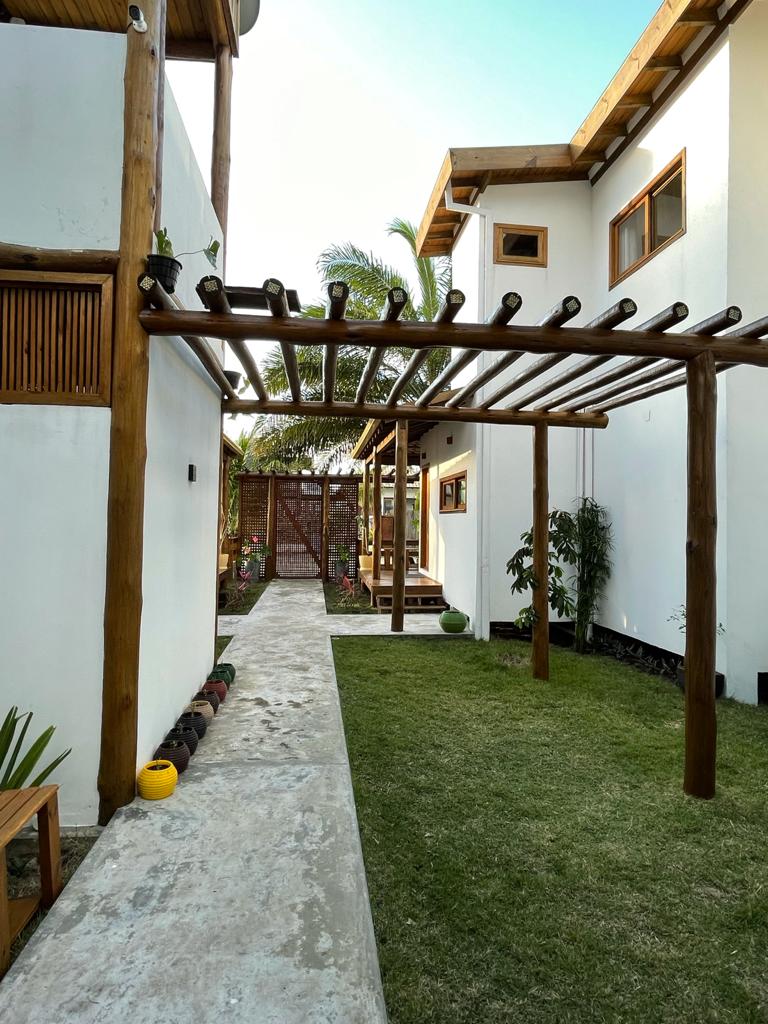 A pathway lined with greenery leads through a well-maintained garden, flanked by houses featuring wooden accents and windows. A pergola with climbing plants creates a shaded area, enhancing the serene atmosphere of the outdoor space.
