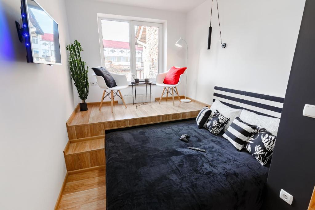 A modern sleeping area features a spacious bed adorned with patterned pillows. Two stylish chairs in bright red are positioned near large windows, allowing natural light to fill the room. A TV is mounted on the wall, while a potted plant adds a touch of greenery.