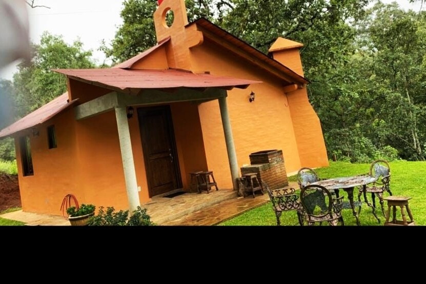 An orange exterior is featured on a cozy structure, complemented by a red roof. A small patio with decorative metal chairs and a table is situated in front, surrounded by lush greenery and trees in the background.