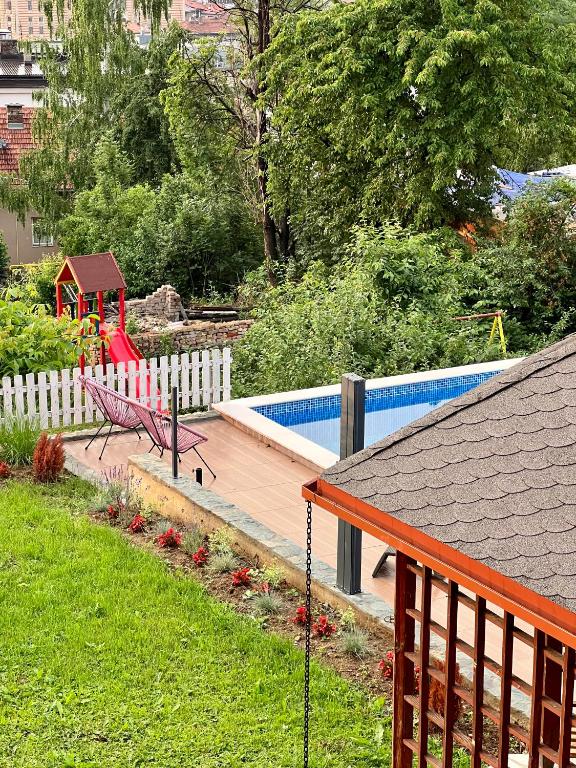 A well-maintained garden area features a swimming pool surrounded by a wooden deck. A hammock is positioned nearby, with trees and a play structure visible in the background. Lush greenery, including blooming flowers, adds to the serene setting.