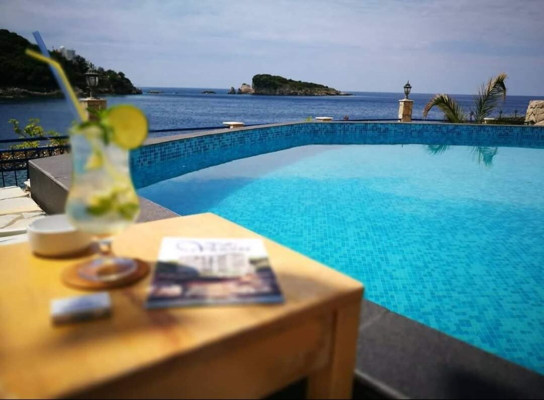 A refreshing drink with a slice of lime is placed on a small wooden table next to a swimming pool. The turquoise water of the pool mirrors the sea beyond, where a distant island is visible under a clear sky. Lush greenery frames the picturesque view.