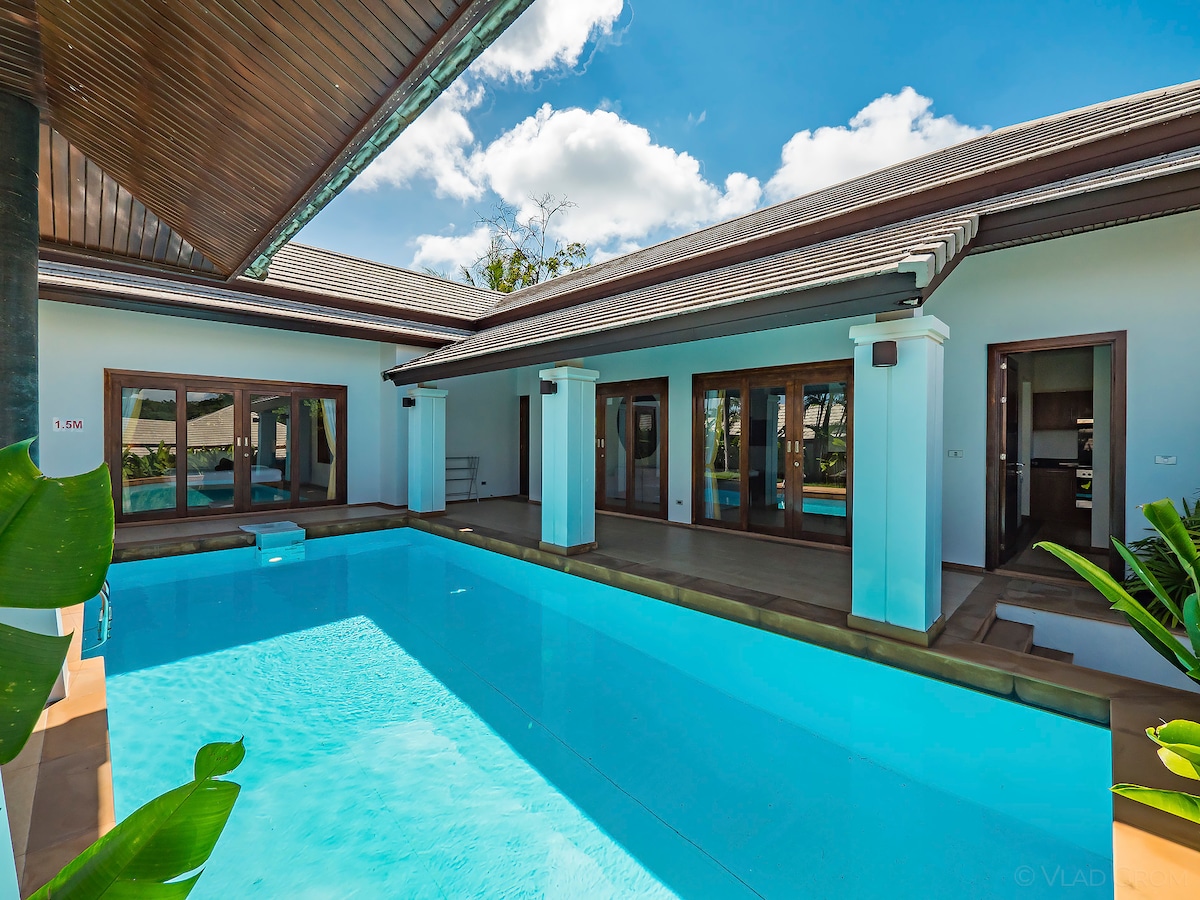 An outdoor view of the villa showcases a private swimming pool surrounded by spacious patio areas. Large glass doors offer a seamless transition between indoor and outdoor living. Lush greenery adds a touch of nature, completing the tranquil atmosphere.