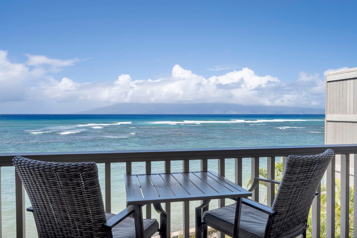 A private lanai features a table and two chairs overlooking the tranquil ocean. The expansive view includes gentle waves and the horizon, with a backdrop of serene clouds against a bright blue sky.