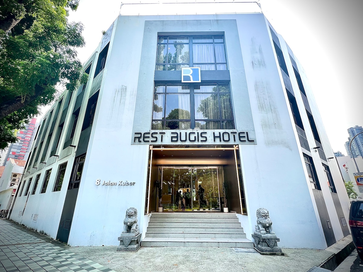 The exterior of Rest Bugis Hotel is showcased, featuring a modern design with large glass windows and a prominent entrance. The hotel name is displayed prominently above the entrance, with surrounding greenery enhancing the contemporary setting.