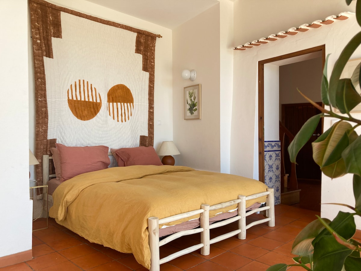 A spacious bed with a warm yellow coverlet is centered in the room, complemented by soft pink pillows. Above, a handmade wall tapestry features simple geometric designs. A lamp and a small plant are placed near the bed, enhancing the serene atmosphere.