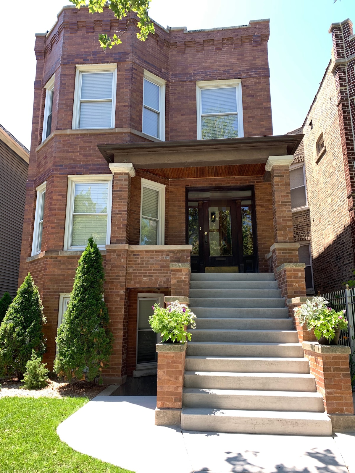 The exterior of a classic Chicago-style brick apartment building is shown, featuring large windows and a welcoming front entrance. A set of steps leads up to the door, flanked by potted plants that add greenery to the space.