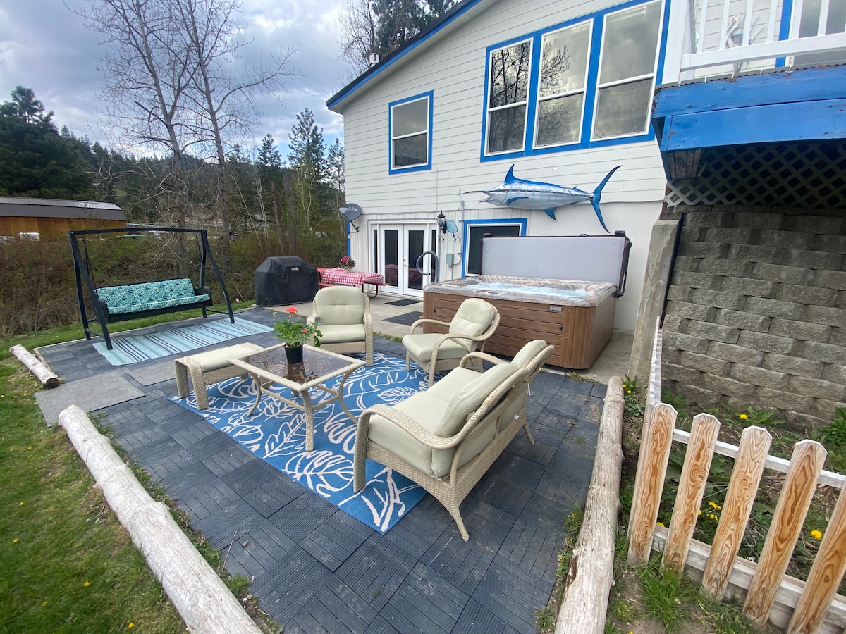 An outdoor seating area features two cream-colored armchairs positioned around a blue-patterned rug. A small table with a floral centerpiece complements a swing and a barbecue grill nearby. The background showcases a large window and a hot tub, surrounded by grass and trees.