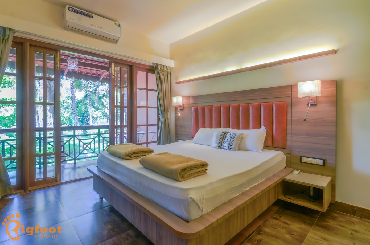 A spacious room features a large bed with crisp linens and decorative pillows. Natural light filters through glass doors that open to a balcony, offering views of greenery. Soft-toned furnishings enhance the calm atmosphere, complemented by practical bedside lighting and air conditioning.