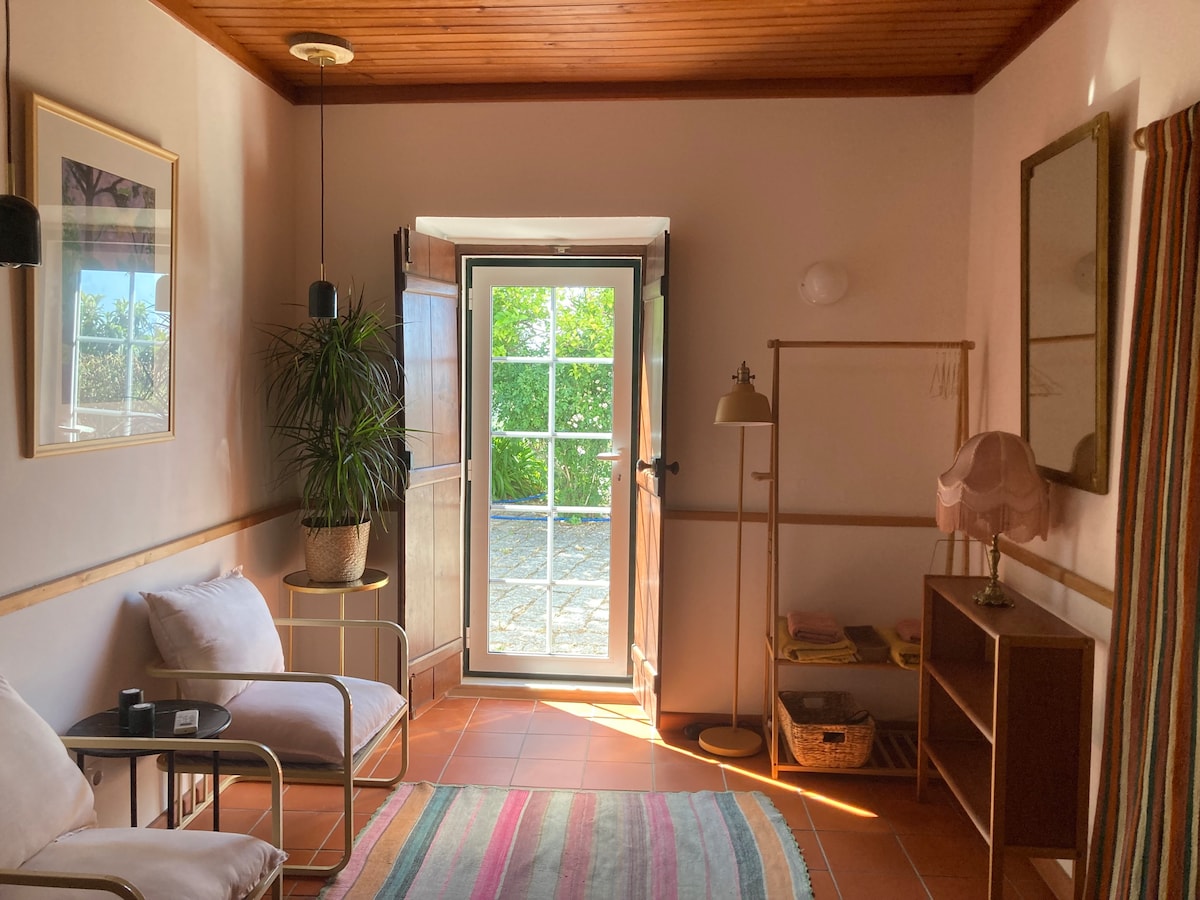 A living area features a comfortable seating arrangement with light-toned chairs and a decorative lamp. A large window allows natural light to fill the space, while a door opens to an outdoor view. Warm-colored walls complement the serene ambiance.