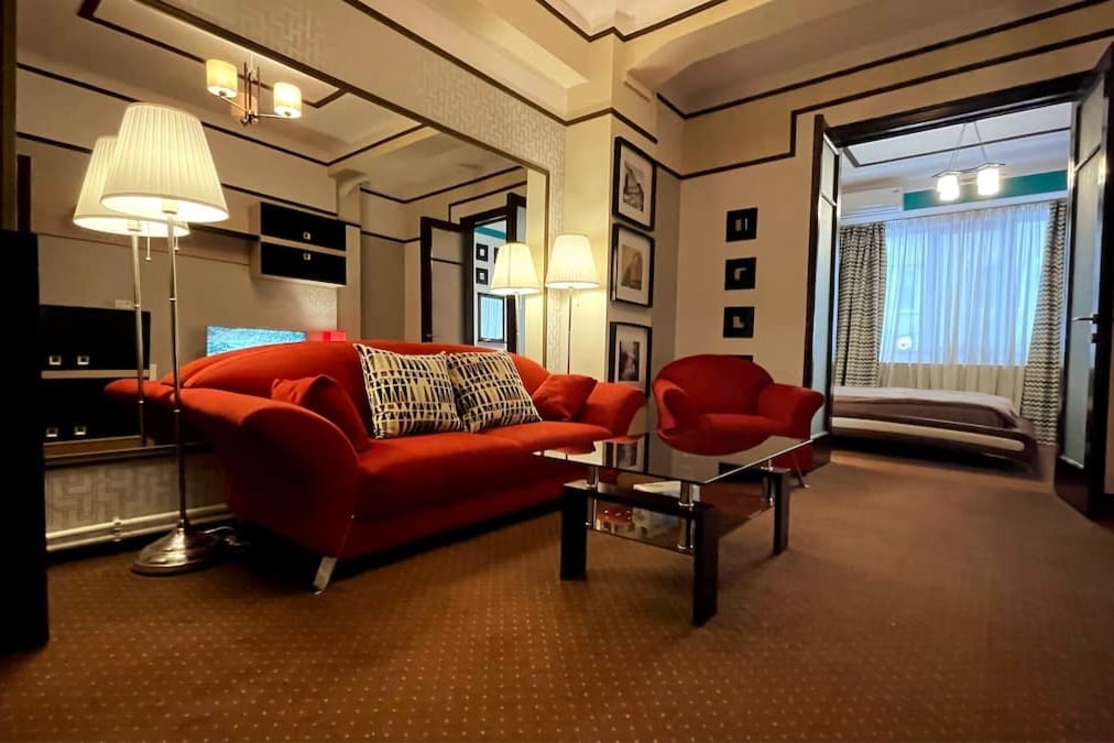 A cozy seating area features a bright red sofa and matching armchairs arranged around a glass coffee table. Warm lighting from stylish floor lamps enhances the decorative elements, while a partially visible bedroom is accessed through an open doorway. The carpeted floor adds to the inviting ambiance.