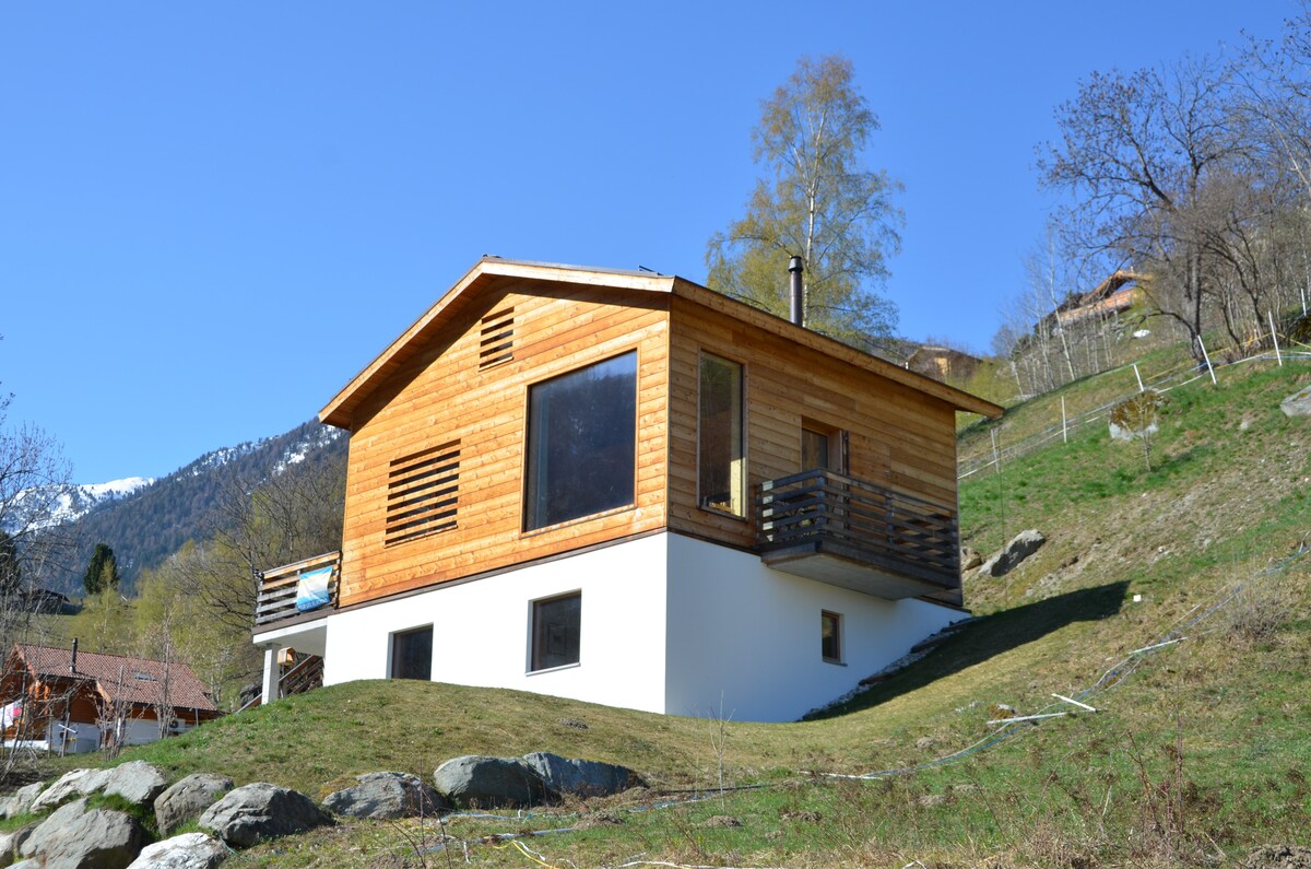A modern chalet features a wooden exterior, blending harmoniously with the natural surroundings. A spacious balcony projects from the upper level, providing views of the landscape. The sloped roof and large windows enhance the structure's contemporary aesthetic while inviting plenty of natural light.
